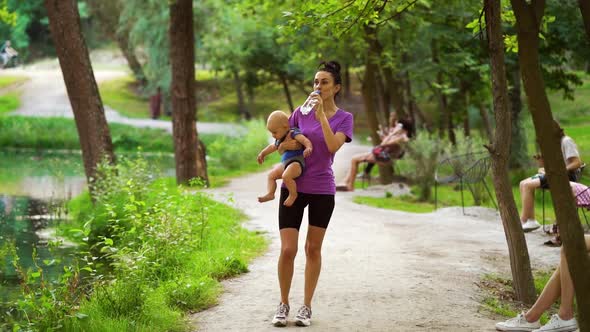 Mom Carrying Baby and Drinking Water After Workout in Park