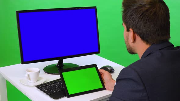 A Young Businessman Sits at a Desk with a Blue Computer Screen and Looks at a Tablet - Green Screen