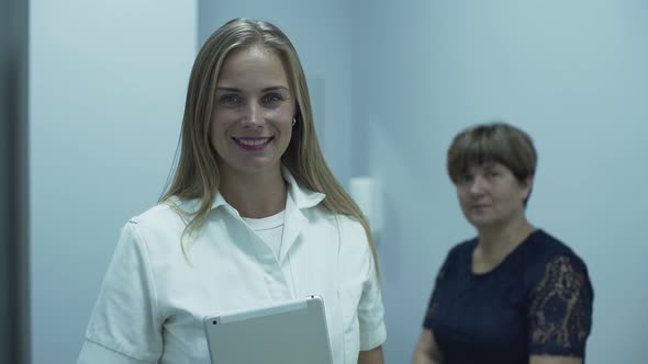 Beautiful Caucasian Doctor Smiling in Foreground Looking at the Camera While Her Mature Patient