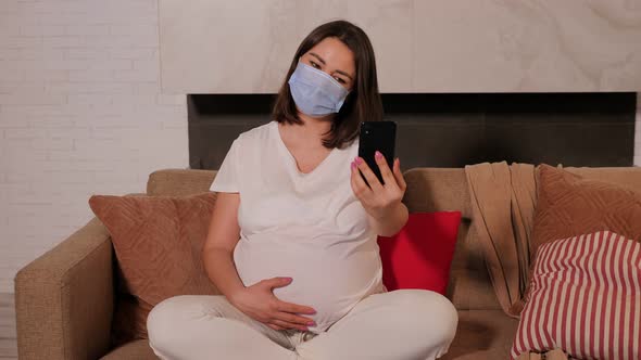 Pregnant Woman in a Mask on the Sofa She Shows a Phone with a Green Screen