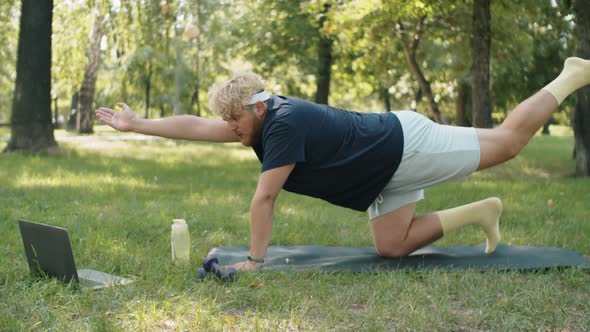 Overweight Man Watching Online Workout on Laptop and Practicing Yoga in Park