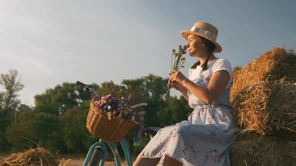 Stylish romantic woman sits on straw near vintage bike and sniffs bouquet of field flowers at sunset
