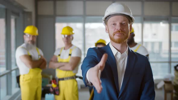 Contractor in Hardhat Stretching Hand for Handshake at Camera with Builders on Background