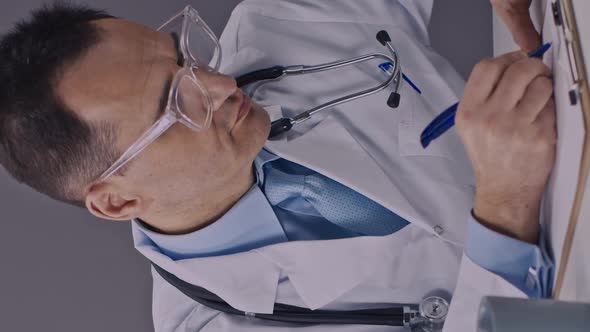 Vertical Shot Focused Doctor Works with Clipboard Writes Medical Record Tired Lays on Desk