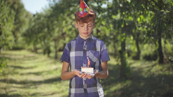 Positive Redhead Teenage Boy Blowing Candle on Birthday Cake Standing in Sunrays in Spring Summer