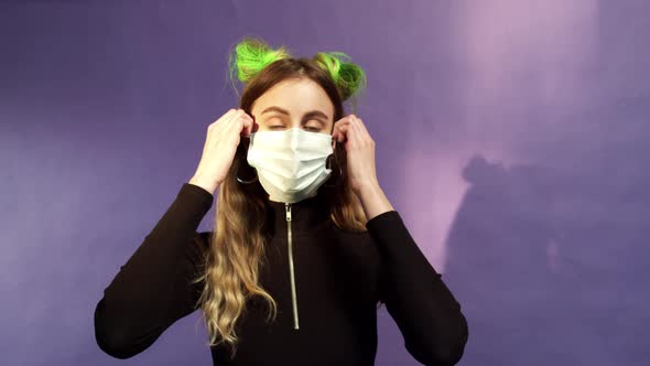 Young Girl with Blonde and Green Hair is Taking Off a Face Mask on a Purple Background