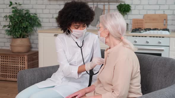 Afro American Woman Doctor in Mask Listens to the Breathing of an Elderly Woman at Home and Supports