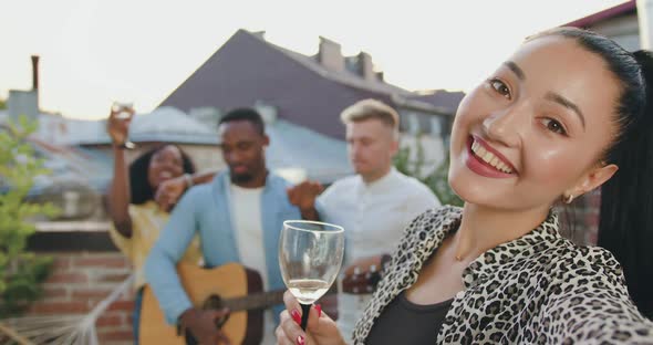 Asian Woman with Glass of Wine Making Selfie on the Background of Friends on Terrace