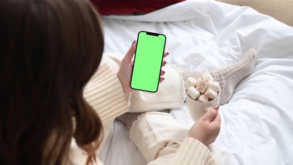 Woman holding modern smartphone with green screen chromakey in hand with cup of cocoa