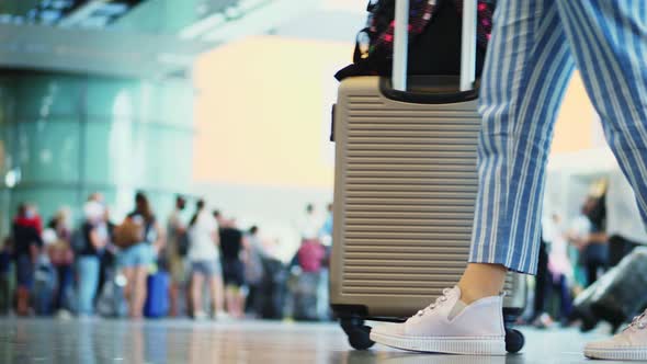 Close-up of Walking Legs in Sneakers, with Suitcase on Wheels, Through Airport Lobby, Against