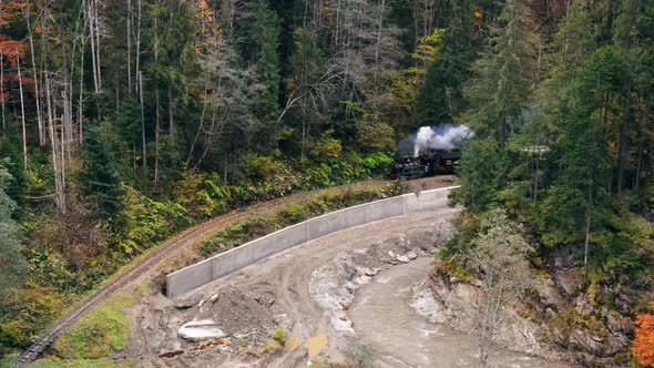 Aerial drone view of the moving steam train Mocanita in a valley along a river, hills covered
