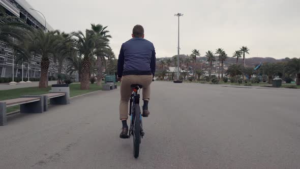 Man Is Riding on Bicycle in Summer Day Over Road in Tropical City Back View