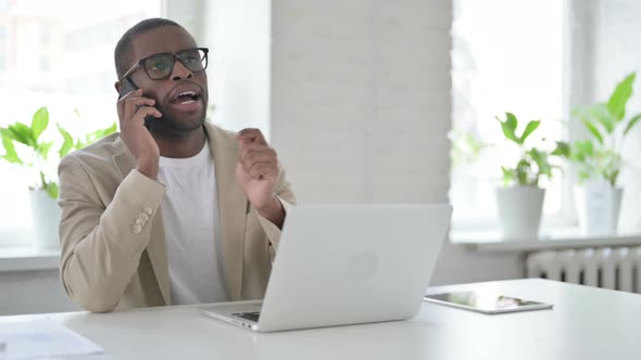 African Man Talking on Smartphone While Using Laptop in Office