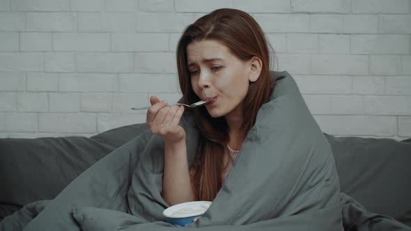 Depressed Woman Eating Ice Cream From Bucket Sitting In Bedroom