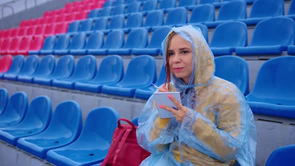 Young Woman in Raincoat with Notepad Pen Sitting on Stadium Bleachers Alone in Rainy Weather