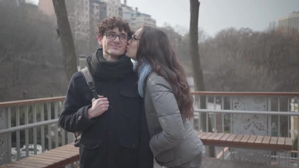 Shy Caucasian Girl in Eyeglasses Kissing Handsome Young Boy on Cheek and Smiling at Camera. Cheerful