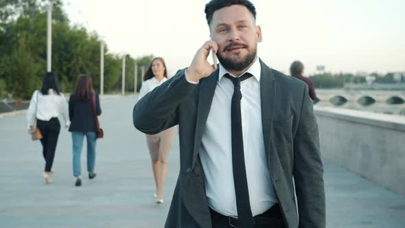 Cheerful Young Businessman Speaking on Mobile Phone Walking Along Embankment in City