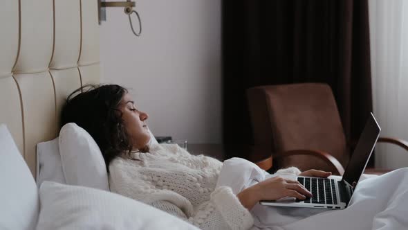 Sleepy Woman Lies on Bed with Laptop in Hands in Modern Apartment