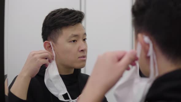 A Young Asian Man Takes Off a Face Mask and Looks at Himself in a Mirror