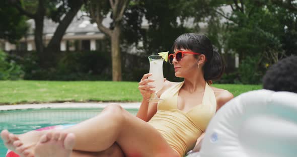 Happy diverse couple wearing swimming suits with inflatables and drink at swimming pool in garden