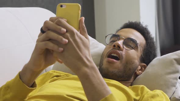 African American Man Lies Resting on the Couch at Home and Holds a Smartphone