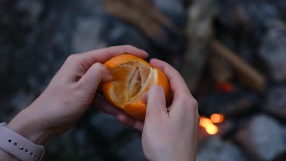 Woman hand peeling ripe sweet tangerine, close up above the fireplace in the forest at camping.