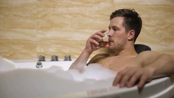 Satisfied Man Drinking Strong Whiskey Lying in Bathtub With Foam, Relaxation