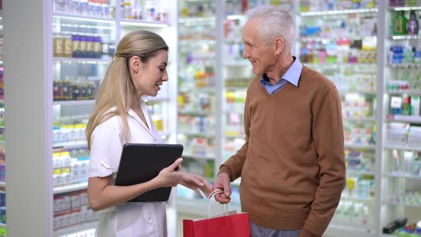 Side View Expert Pharmacist Passing Shopping Bag with Medications to Senior Client Leaving Drugstore