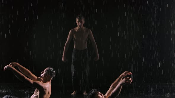 A Group of Three Acrobats Perform Difficult Balancing Tricks in the Rain. Men Stand Against Black