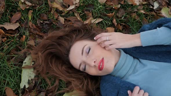 Young Redhaired Woman in a Blue Coat Lies on the Grass in an Autumn Park Leaves Lie Around