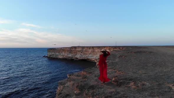 Aerial View of a Young Woman in a Red Dress and a Straw Hat Walks Along the Edge Front of a Cliff By