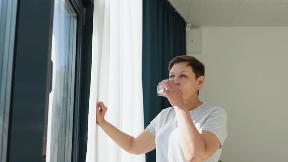 Senior Woman Drinks Water Standing Near the Window in a Light Room