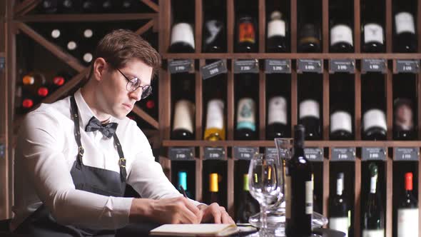 Male Wine Steward Looks at Bottles of Wine and Writing in Notebook at Wine Store