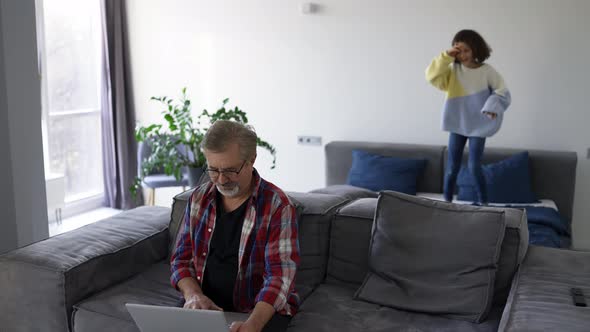 Adorable Little Girl Playing on the Background While Grandfather Using Laptop at Home