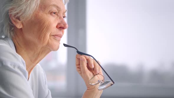 Thoughtful aged woman in blue blouse holds designed glasses