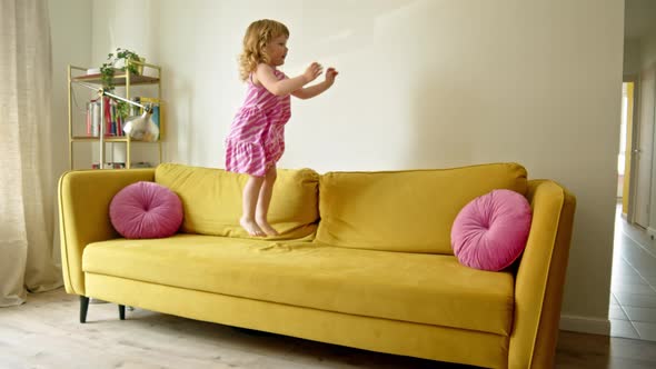 Cheerful and Cute Preschool Girl Jumps and Dances on Yellow Large Sofa in Living Room