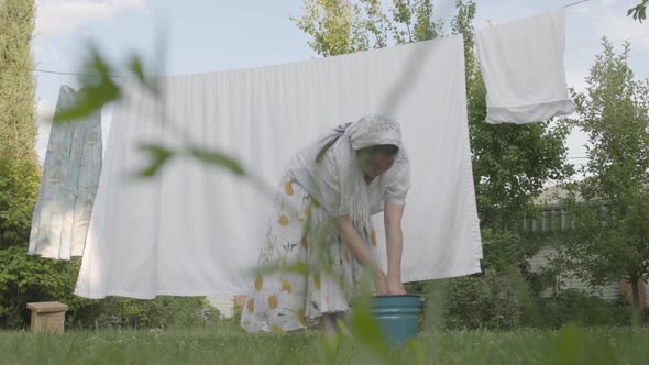 Attractive Senior Woman with a White Shawl on Her Head Hanging Bed Linen on the Rope in the Garden