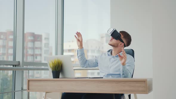 A Young Man Sitting at a Desk in the Office Uses Augmented Reality Glasses To Work on Business