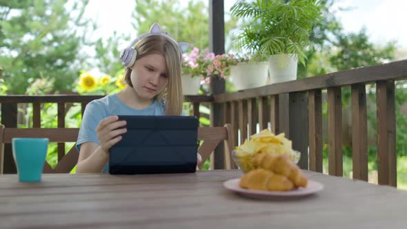 Teenage Girl Sits At Wooden Table In a Summer Cafe In Headphones With Phone