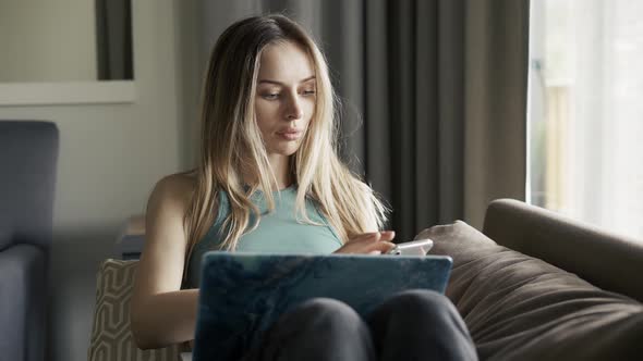 Blonde Woman Sitting on Sofa with Laptop on Knees Using Her Cell Phone
