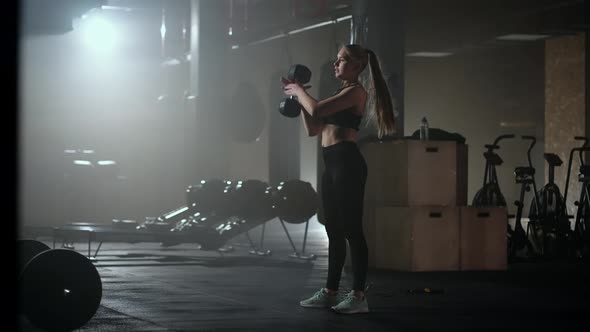 A Strong Woman Makes Efforts and Overcoming Difficulties Lifts a Dumbbell in a Dark Gym