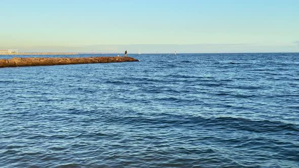 A man is standing on a rock wall fishing in the ocean at sunset.