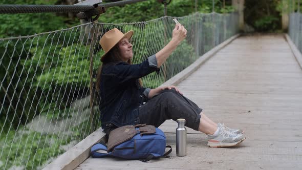 Pretty woman traveler in hat taking photo with phone while sitting on a suspended bridge