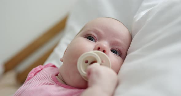 Medium Shot of Adorable Sleepy Female Infant Wearing Ping T-shirt Lying on Bed and Sucking White