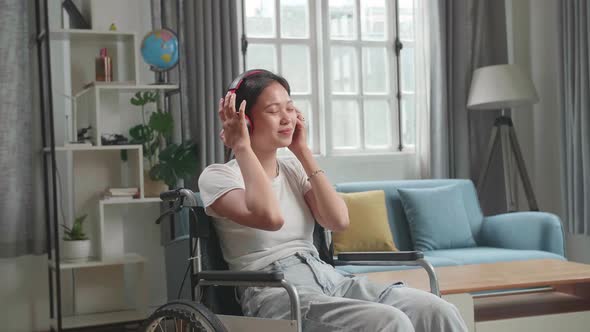 Asian Woman Sitting In A Wheelchair Listening To Music With Headphones In Living Room