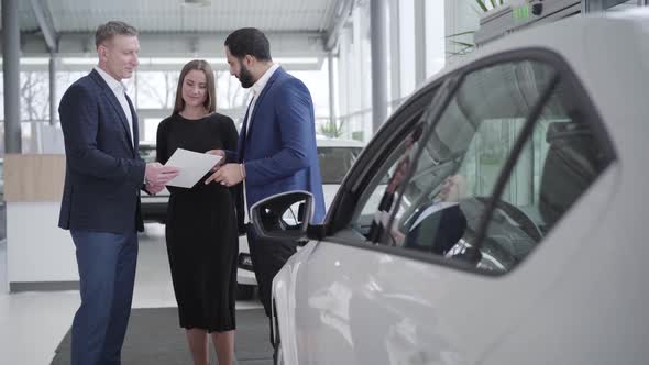 Adult Successful Family Talking with Car Dealer in Showroom. Young Confident Middle Eastern Husband