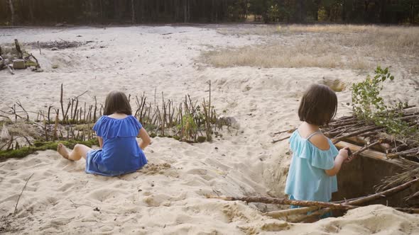 Two Little Girls in Colorful Dresses Are Playing on the Sand in the Forest 