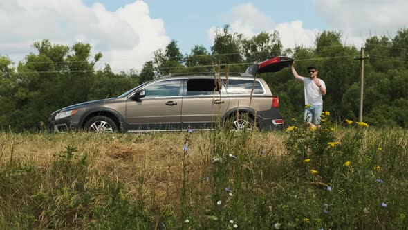 Man in White Tshirt and Black Cap with Sunglasses Stands Near Open Trunk of Brown Station Wagon on