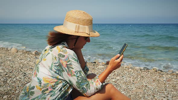 Young female in straw hat and summer tropical shirt sitting on beach, relaxing and using smartphone.