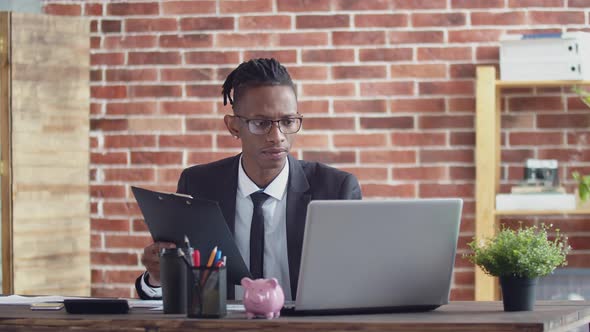 Black Man Businessman in Glasses and a Suit Sits at an Office Table and Enters Information From a